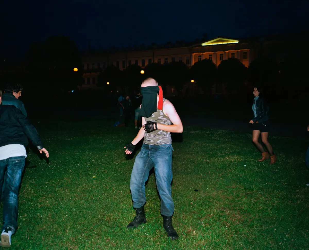 Masked person wearing camouflage tank top and blue jeans runs across a grassy park at night, with a lit building in the background.