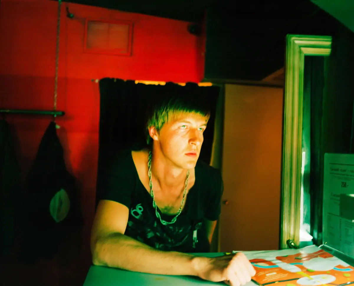 Person behind a bar counter, leaning forward with a chain necklace, under red and green neon lighting in a dim room.