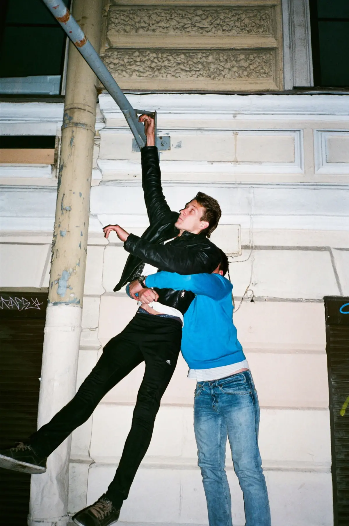 Two people on an urban building exterior; one wearing a black jacket climbs a rusted pipe, supported by the other in a blue hoodie.