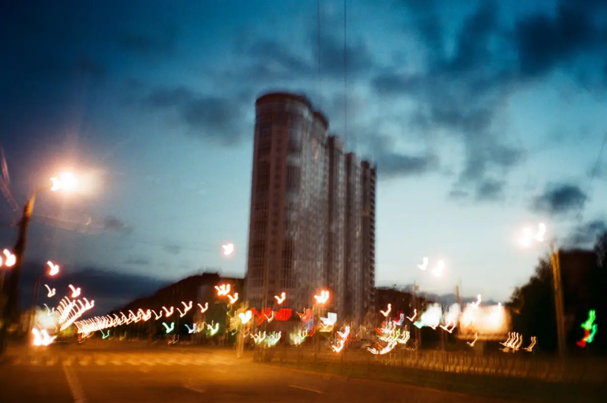 Blurry urban night scene featuring a tall cylindrical high-rise, with streaking streetlights and a cloudy sky.