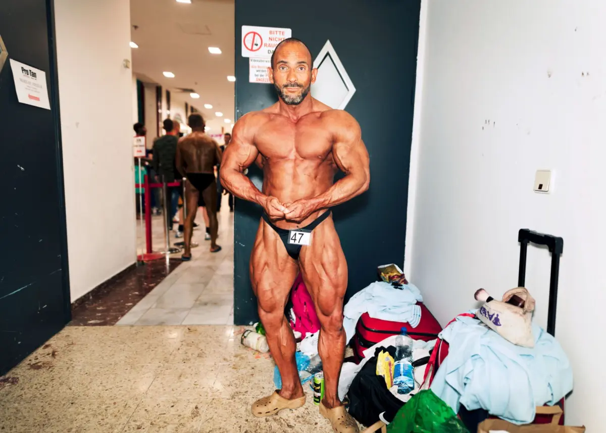 Muscular bodybuilder flexing in doorway backstage at a competition; other contestants and scattered gear in the hallway.