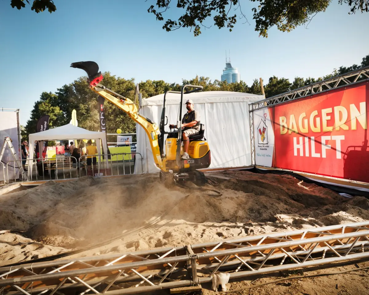 Operator on a yellow mini excavator lifts a sand pile at an outdoor exhibition with white tents and a red banner.