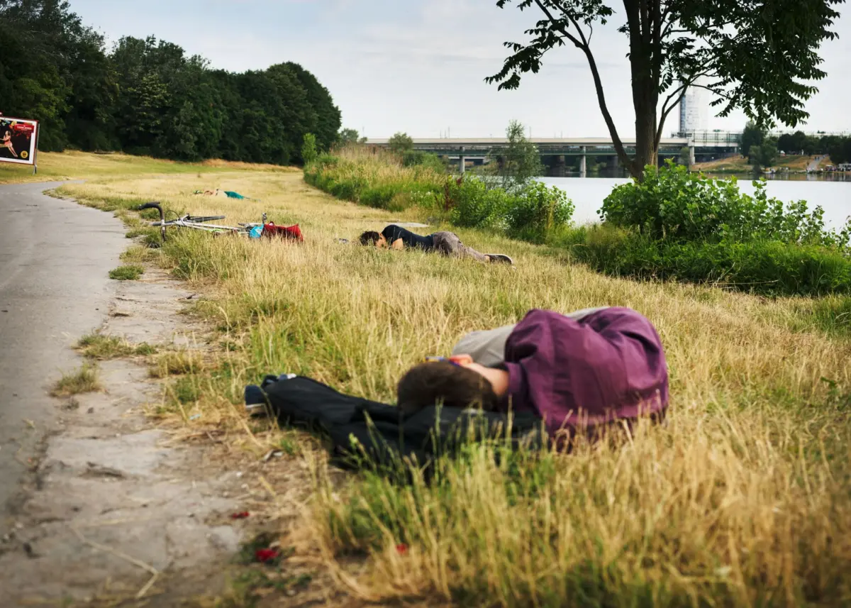 People lying on a grassy riverbank beside a paved path; a bicycle rests on the path with a bridge across the river in the distance.
