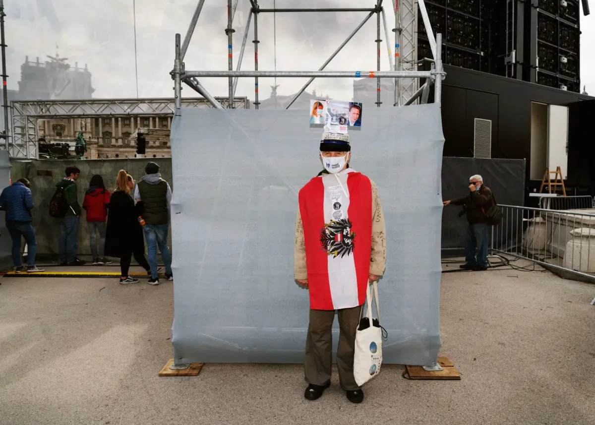 Person in red-and-white robe and hat with taped photos stands before a gray fabric barrier at an outdoor stage with scaffolding.