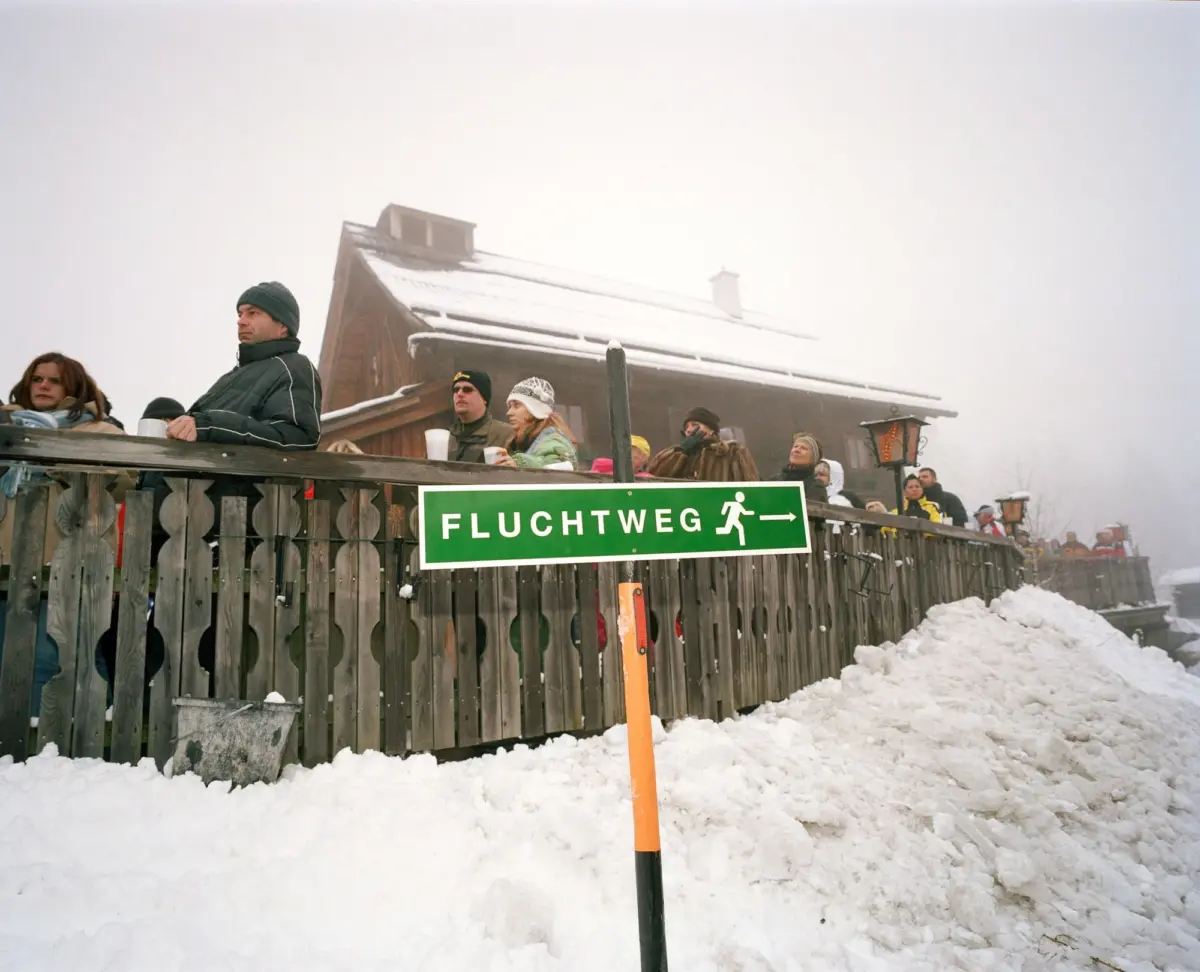 Group of winter-clad spectators on a wooden balcony above snow, holding cups; foreground green 'FLUCHTWEG' sign near a snow-dusted chalet.