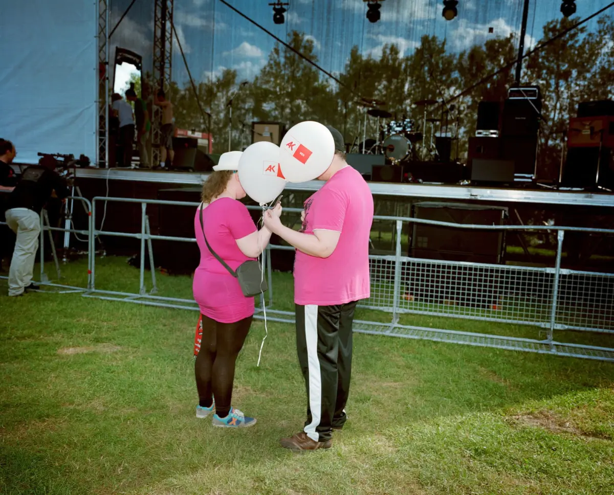 Two people in pink shirts kiss while holding white balloons in front of an outdoor concert stage.