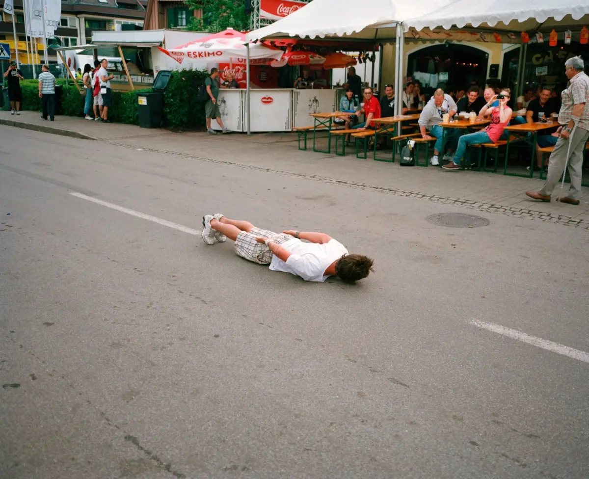 Two people lying prone on a street during a festival, with food stalls and people seated under white tents in the background.
