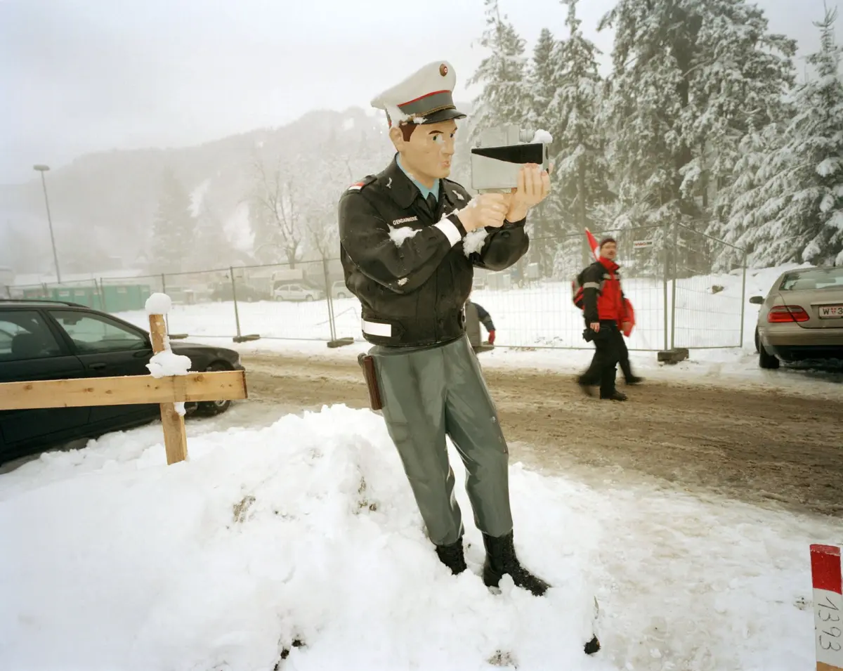 Life-size gendarmerie officer statue aiming a handheld speed gun in a snowy outdoor parking lot.