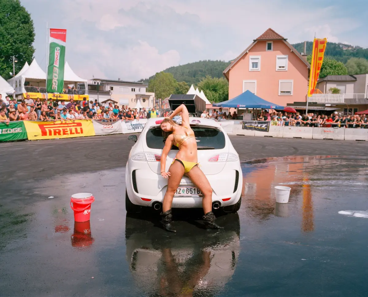 Bikini-clad person poses on the rear of a white car on wet pavement at a crowded street event, with banners and tents in the background.