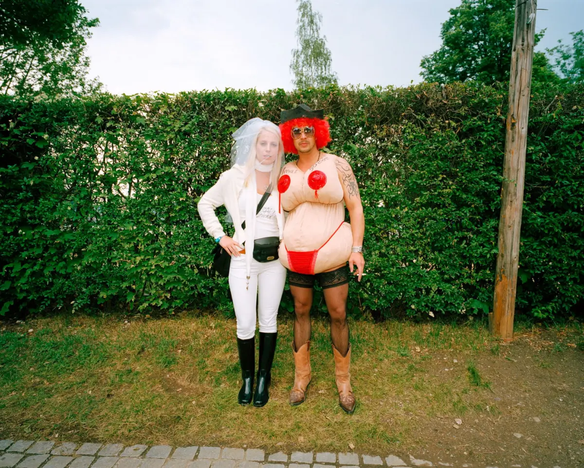Two adults in costumes pose outdoors by a hedge; left in white bridal outfit with veil, right in red wig with sunglasses and inflatable torso costume.