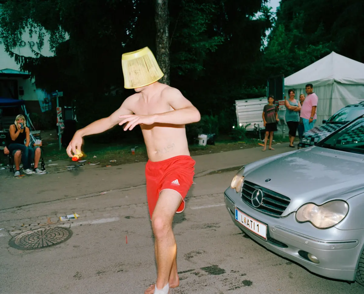 Shirtless man in red shorts dances on a street with a yellow lampshade on his head, near a white tent and spectators.