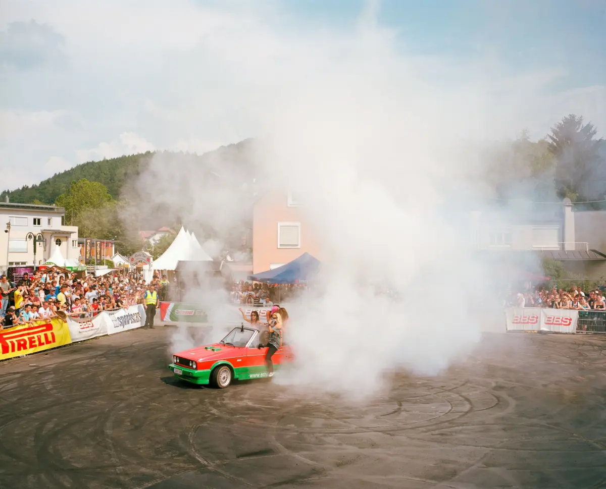 Red and green vintage rally car performing a burnout amid billowing smoke as crowd watches from barriers at a street festival.