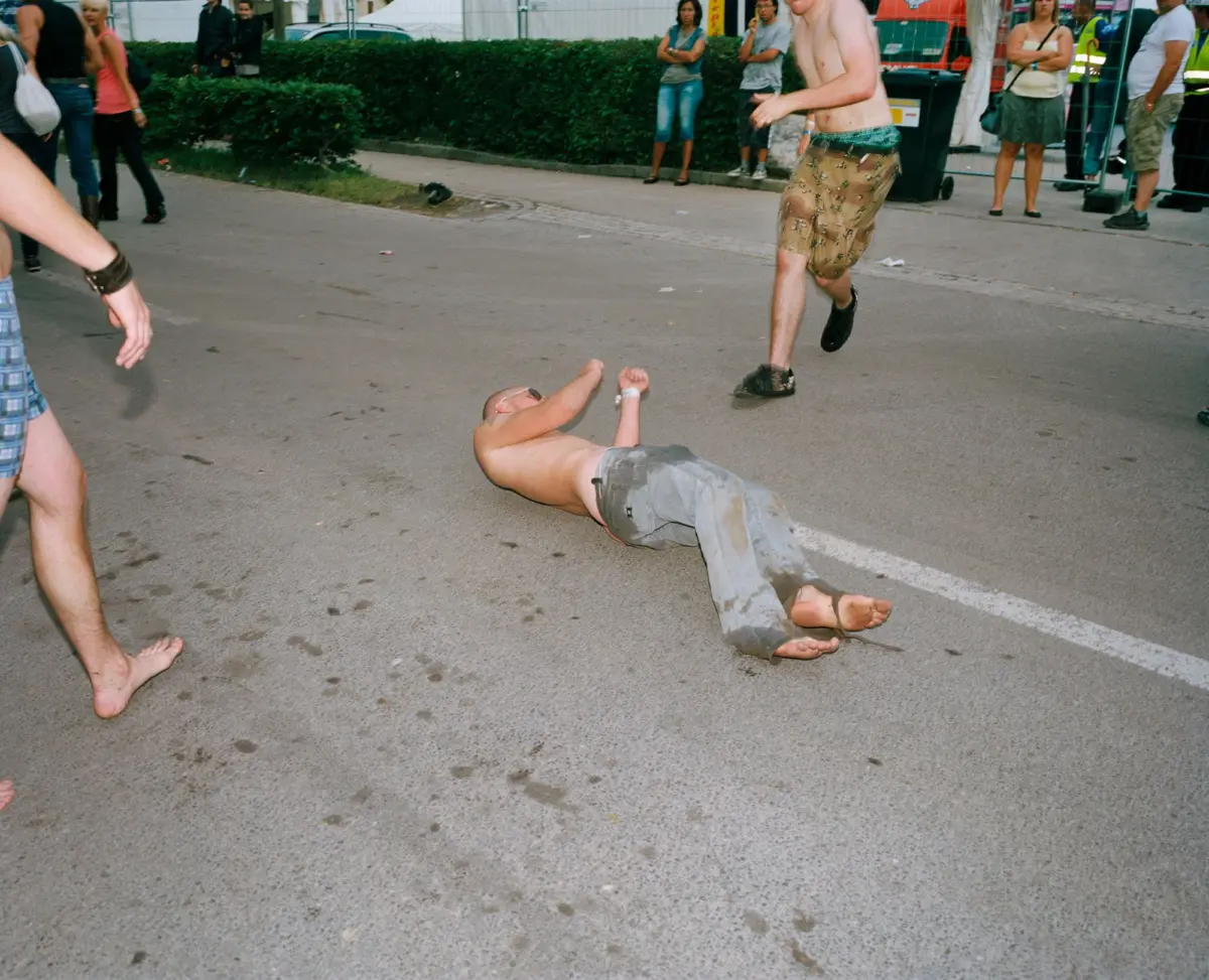 Two shirtless men on a street; one stands while the other lies on the pavement as bystanders watch.