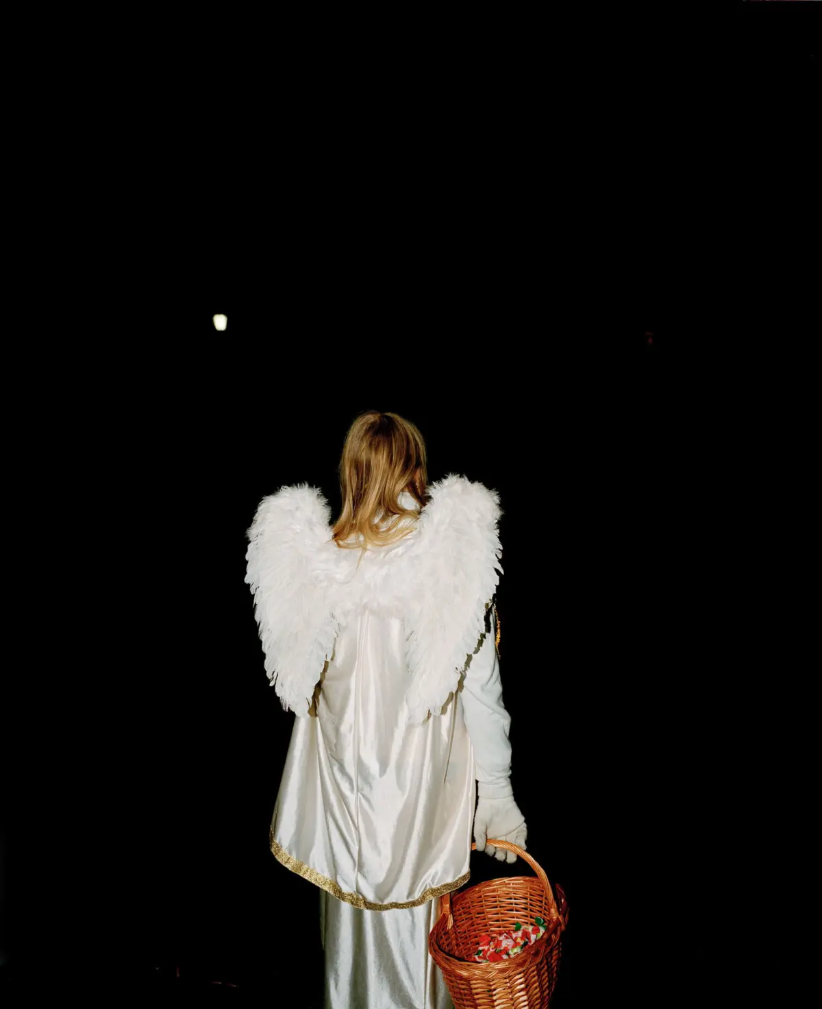 Back view of a person in a white angel costume with feathered wings, standing on a dark stage and holding a wicker basket.