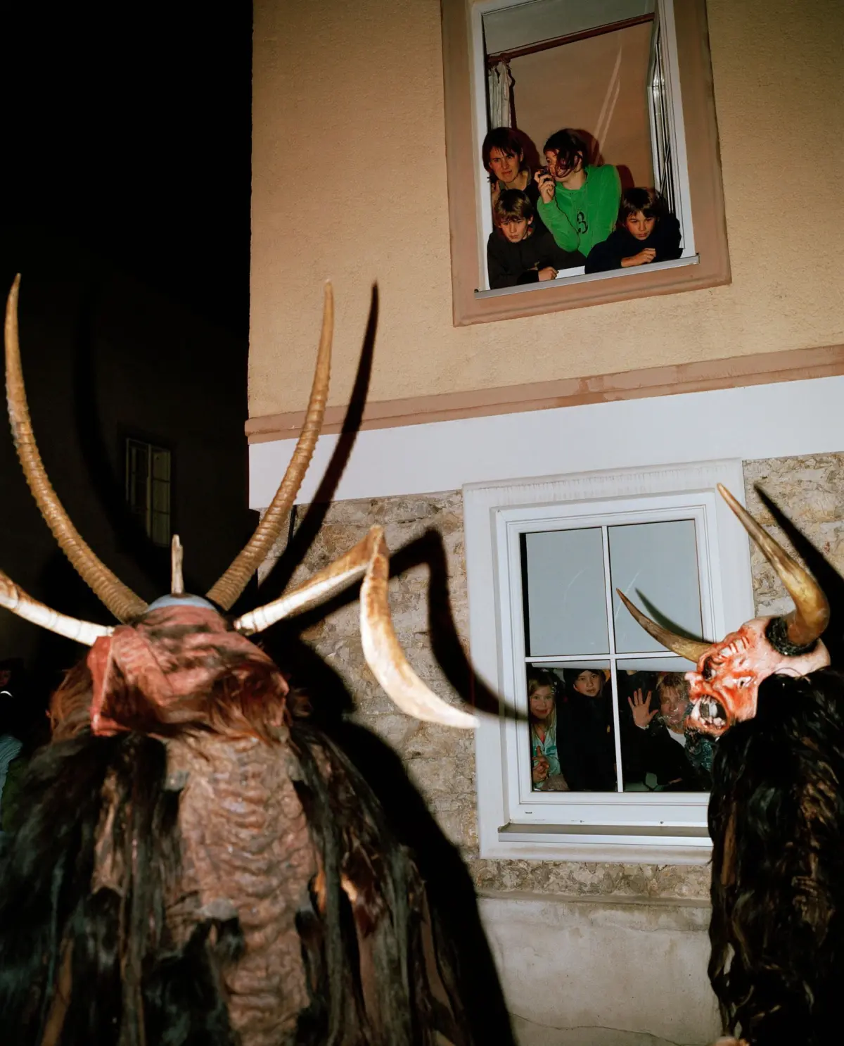 Demon-costumed figures with long horns loom outside a beige house while a family watches from an open window at night.