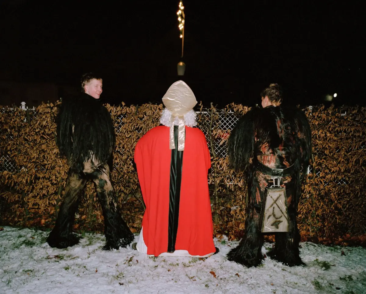 Three costumed people outdoors at night; central figure in red robe with white headpiece, flanked by two in black feathered cloaks; snowy ground.