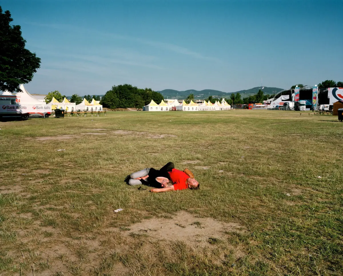 Person in red shirt lying on grass at an outdoor festival with white and yellow-topped tents and a distant stage.