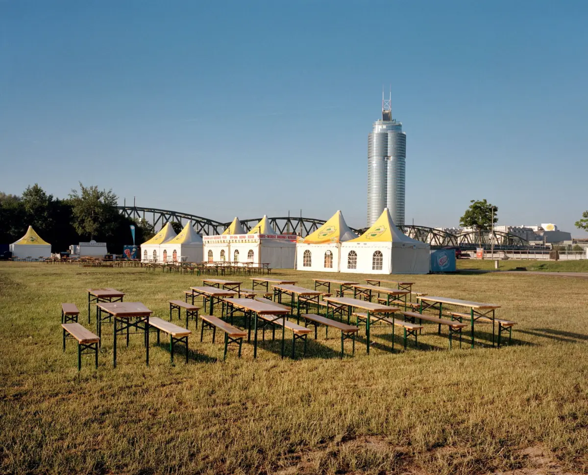 Outdoor festival setup with white tents and wooden picnic tables on a grassy field; a tall glass skyscraper and bridge in the background.