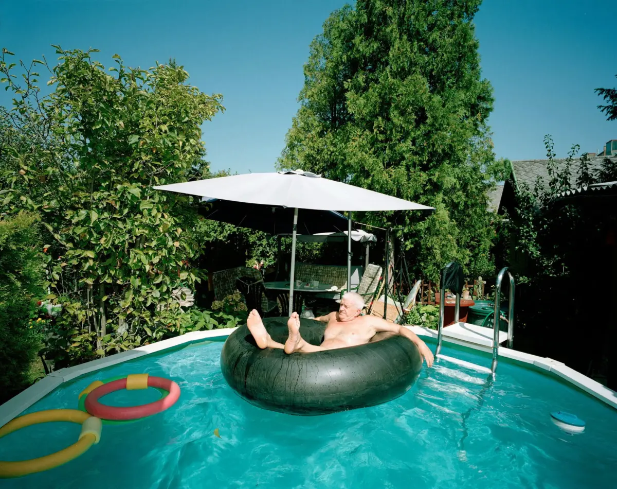 Older person lounging on a black inflatable inner tube in a backyard pool, beneath a large white umbrella, with lush trees surrounding.