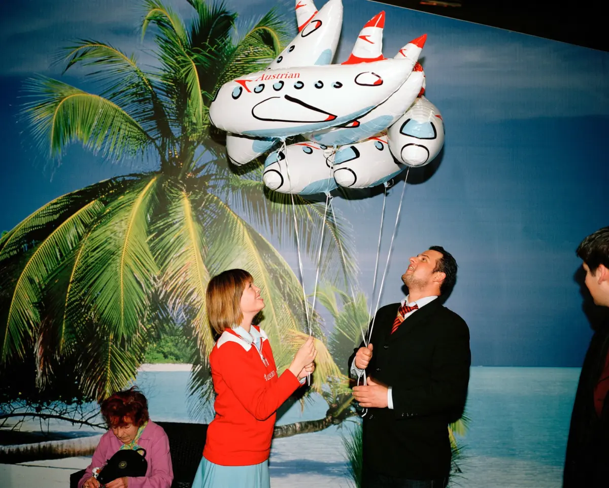 Two adults hold strings of a large white inflatable ship balloon against a tropical palm backdrop.