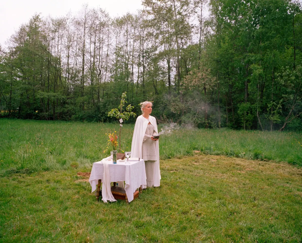 Woman in a white robe stands beside a table set with flowers and glassware in a grassy clearing, a forest backdrop.
