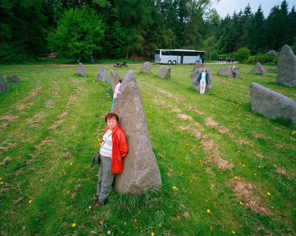 Person in a red jacket leaning against a tall standing stone in a grassy park with a ring of stones and a white bus in the background.