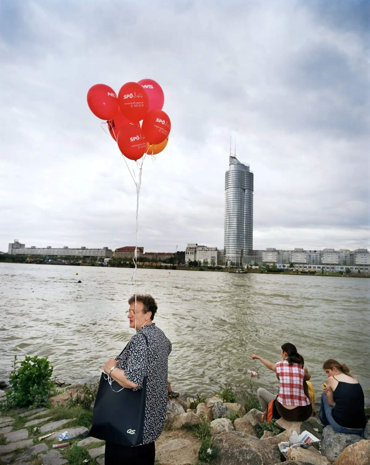 Cluster of red balloons rising over a river with a city skyline; foreground shows a woman with a black tote standing on a rocky riverside.