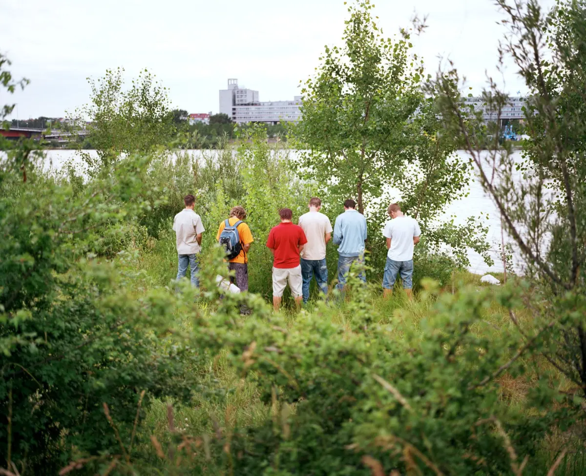 Group of people standing with backs to the camera on a grassy riverside, facing water and distant city buildings.