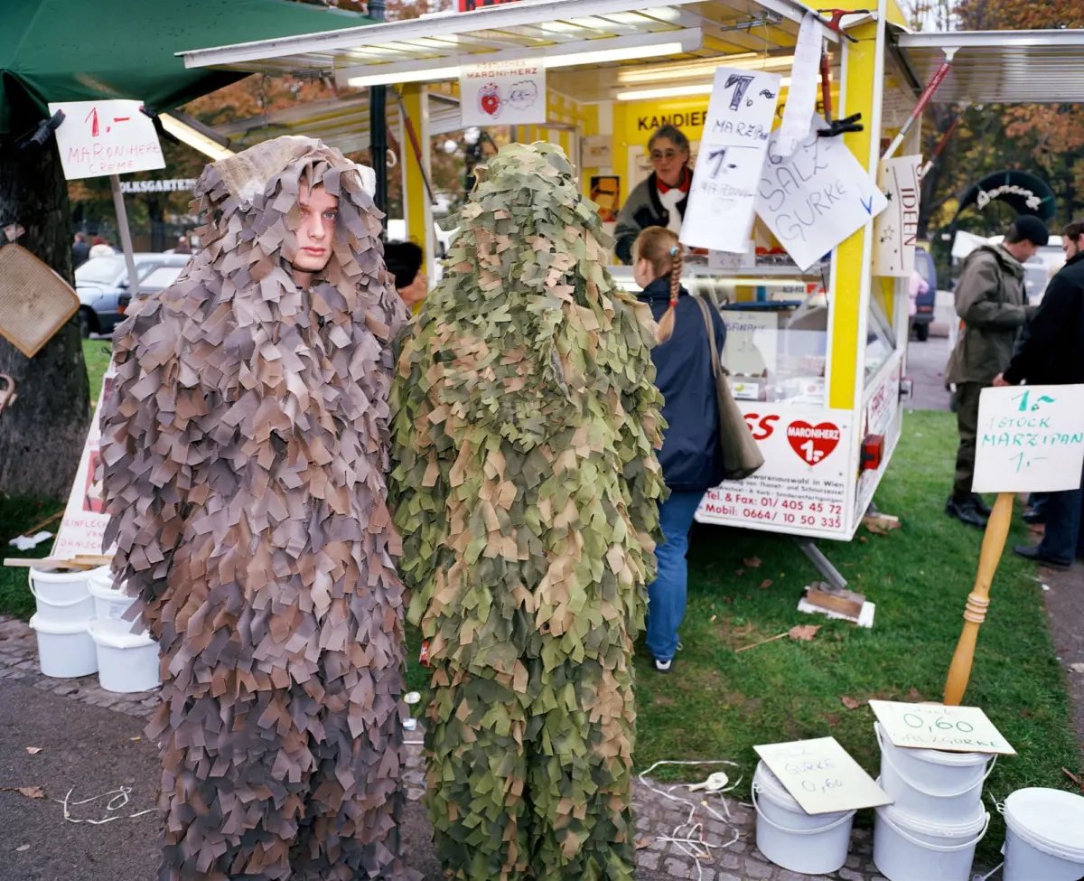 Two people wearing camouflage ghillie suits stand at a yellow outdoor market stall.