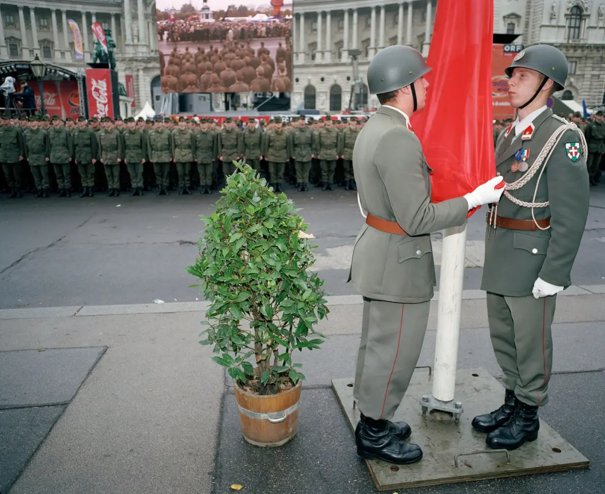 Two soldiers present colors with a red flag at a military ceremony; rows of troops and a historic building in the background.