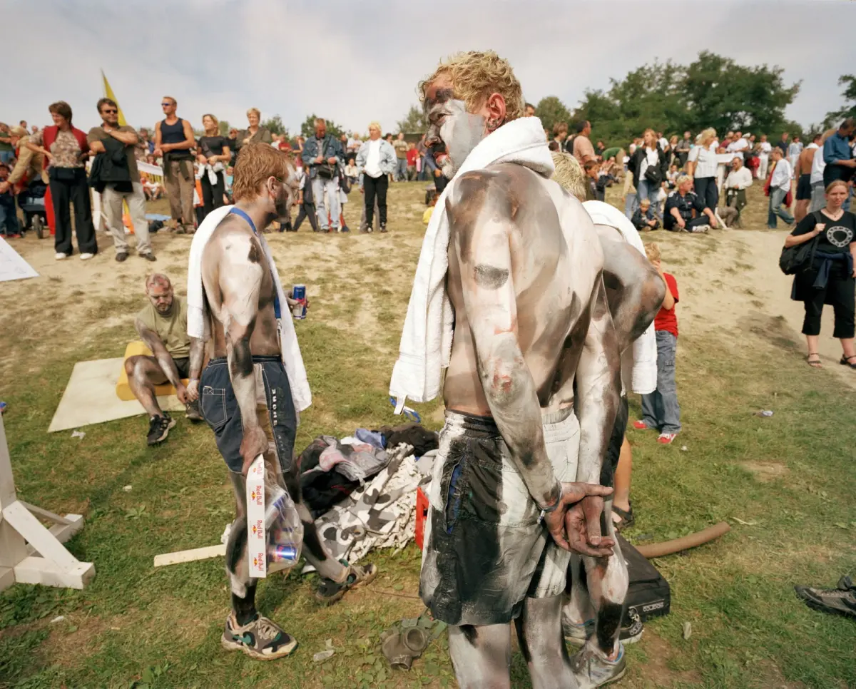 Two shirtless people with white and black body paint and towels around their shoulders stand in a grassy field at a crowded outdoor festival.