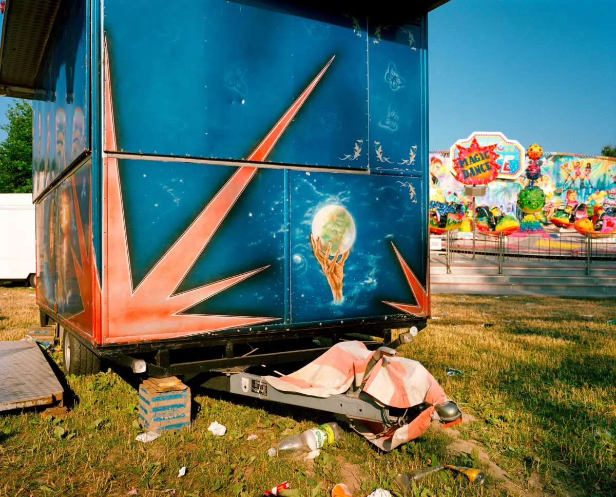 Blue space-themed carnival trailer on grassy lot; scattered litter around, colorful fair rides in the background.
