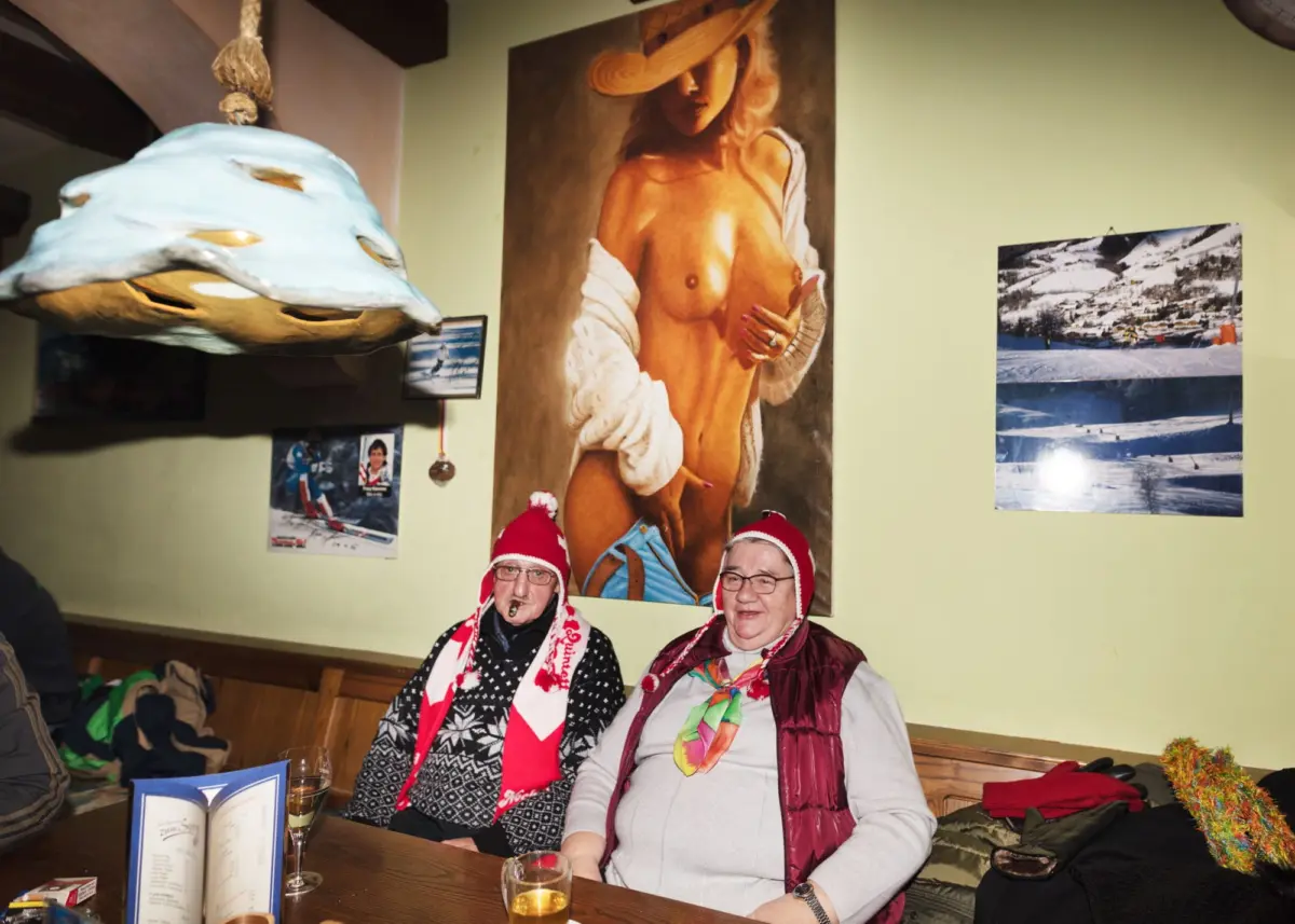 Two elderly women in knit hats and scarves sit at a wooden table, posing in a cozy room with wall art: a nude painting and a snowy landscape photo.