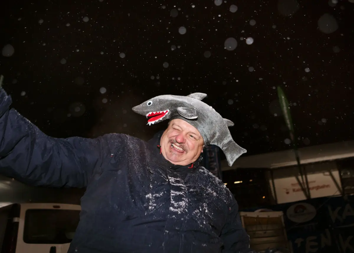 Adult person wearing a plush gray shark hat, arms raised, smiling as snow falls at night in a parking lot.