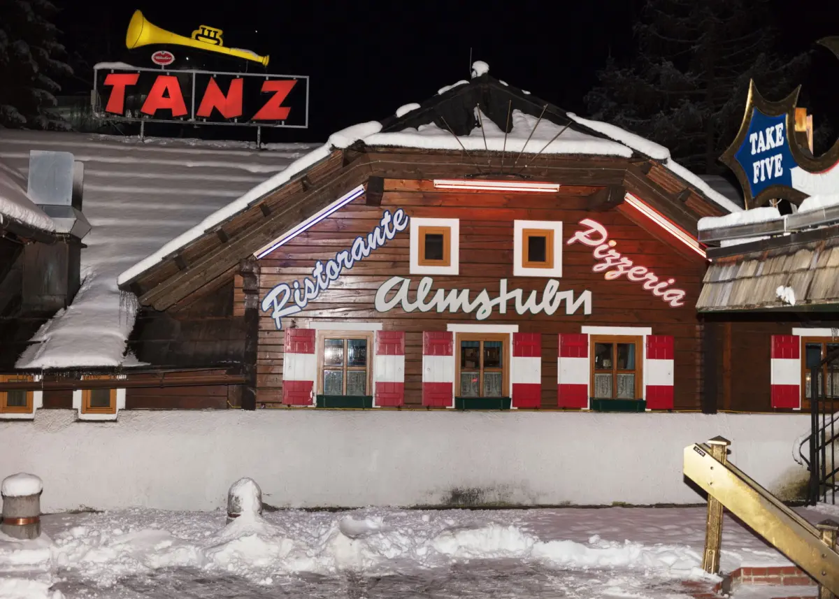 Alpine wooden restaurant at night with snow on the roof, neon lights; large red TANZ sign and blue Take Five sign nearby.