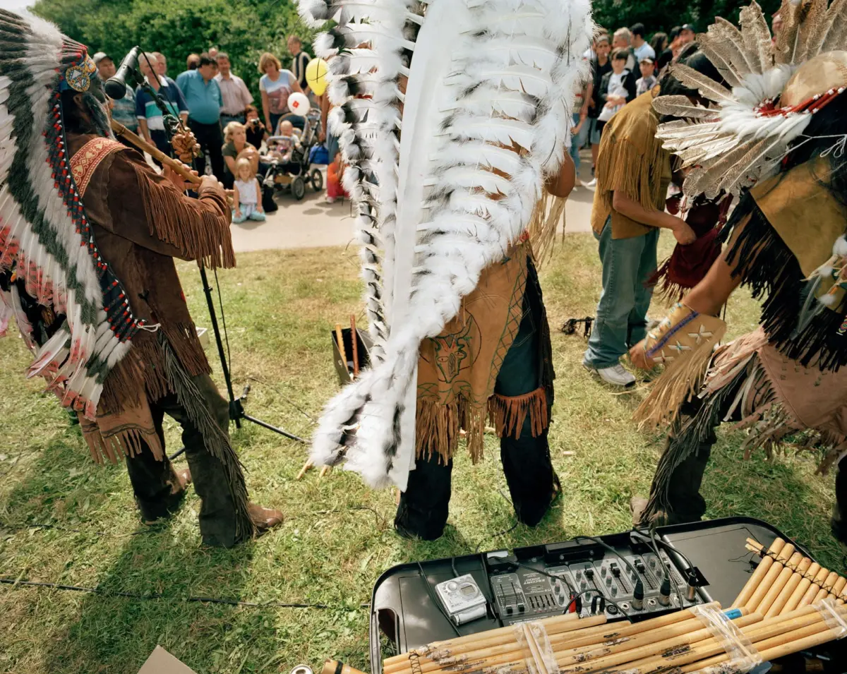 Indigenous dancers in feathered regalia perform outdoors; crowd watches in a park, with audio equipment visible in the foreground.