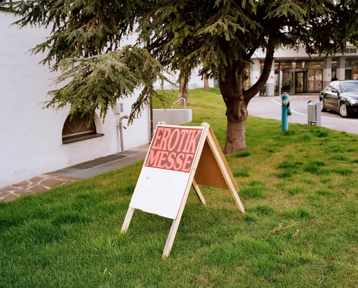 Large evergreen tree overhangs an A-frame sign on a grassy patch beside a sidewalk, with a storefront and parked car in the background.