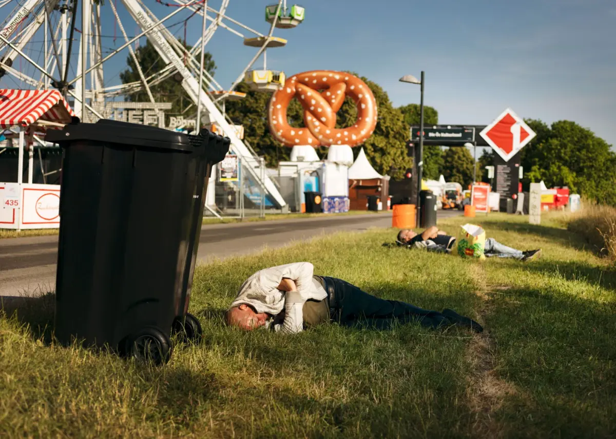 Person lying on grass beside a large black trash bin at a fairground with a Ferris wheel and oversized pretzel sculpture.