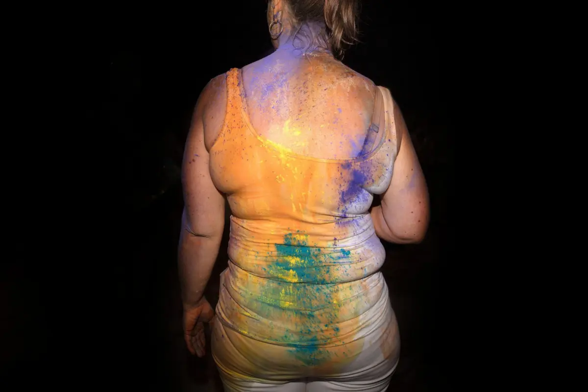 Back view of an adult person in an orange tank top and white pants, covered in blue, yellow, orange, and purple powder at a color festival.