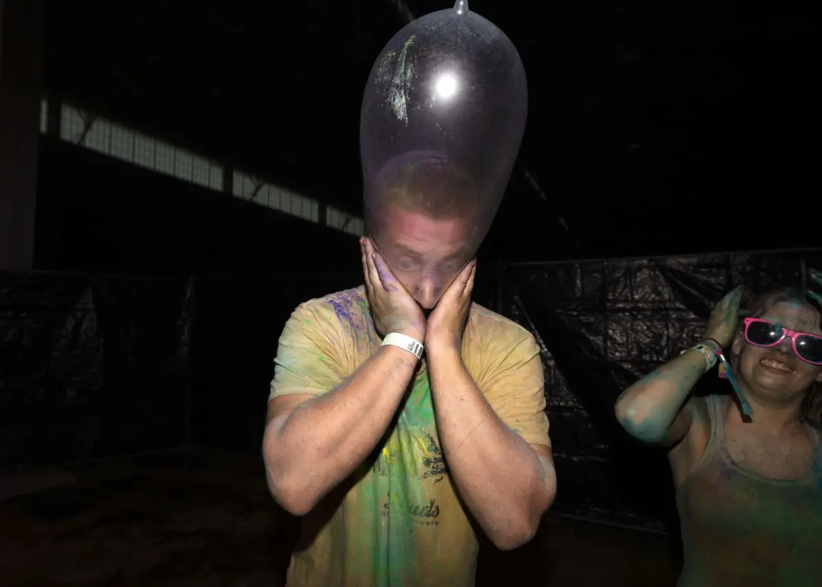 Person holds a large balloon on their head, hands on cheeks, paint-splattered shirt, in a dark party setting; nearby person wears pink sunglasses.