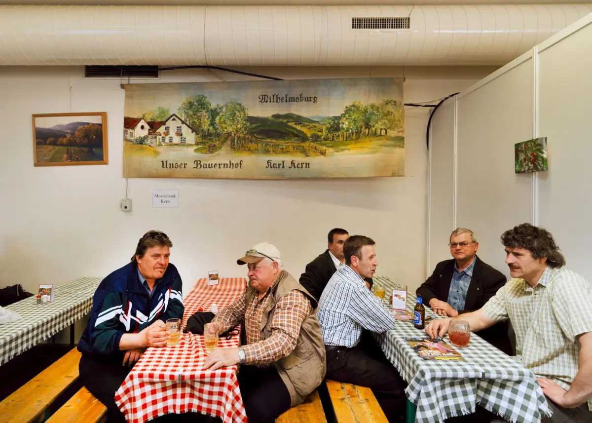 Group of men seated at long benches, talking and drinking indoors; red-and-white checkered tables; banner reads Wilhelmsburg Unser Bauernhof Karl Kern.