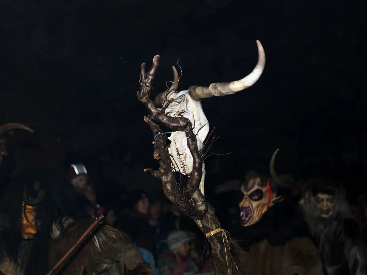 Nighttime ritual with a figure holding a staff topped by a white bull skull and curved horn; masked participants in the dark.