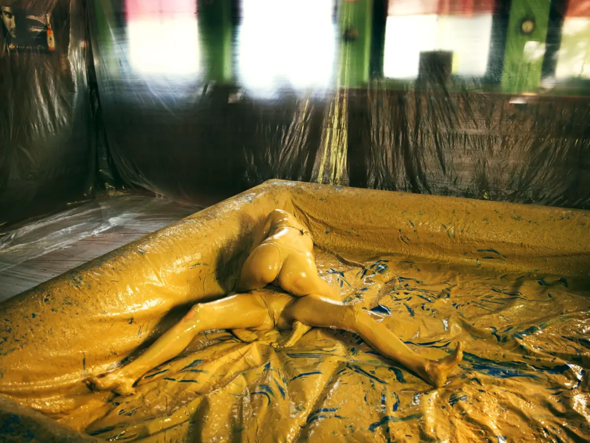 Person in yellow protective suit covered in yellow slime lying on a slime-covered floor inside a plastic-walled indoor space.