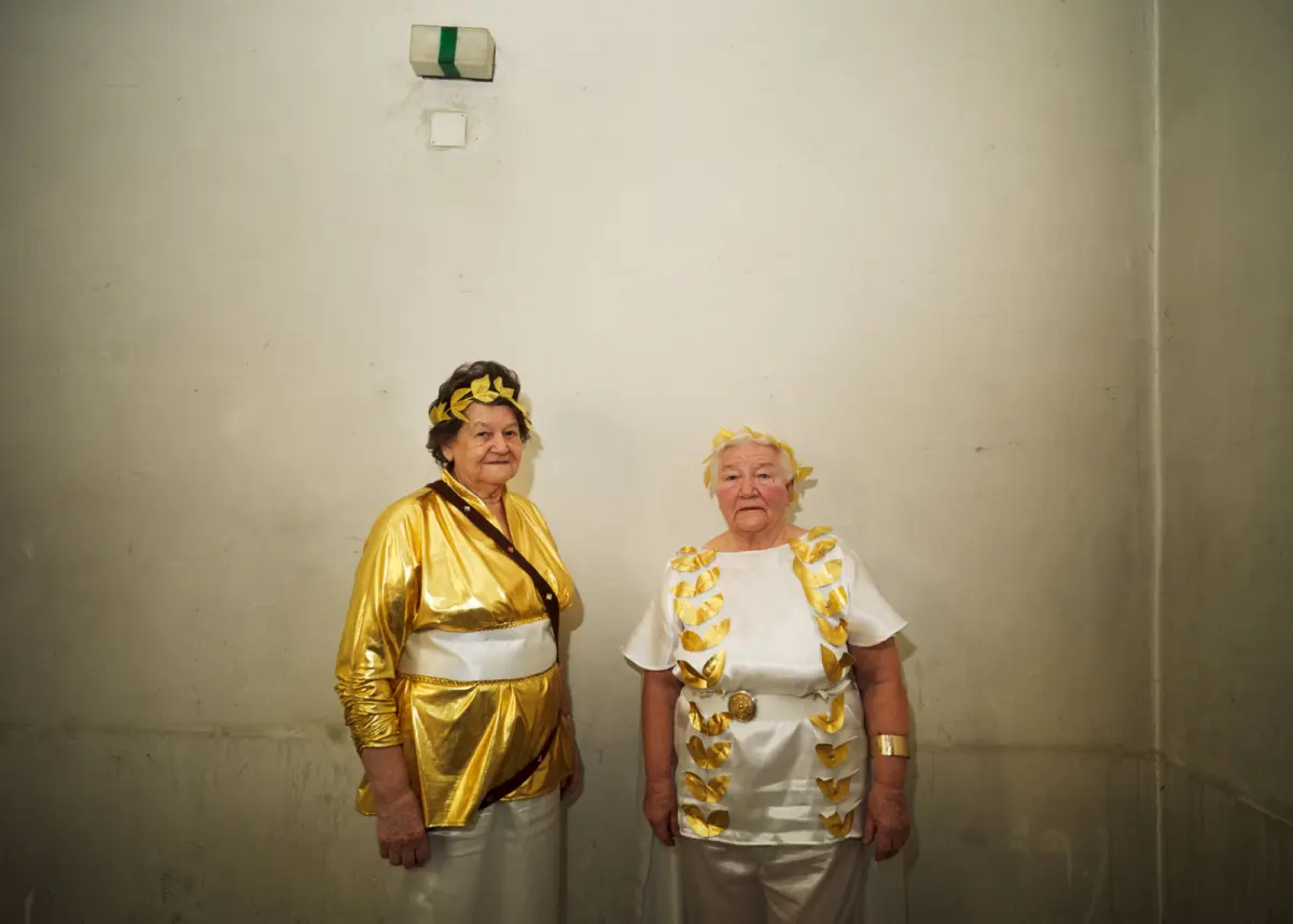 Two elderly women in gold and white Greek-inspired outfits with laurel wreaths, standing and posing against a plain beige wall.