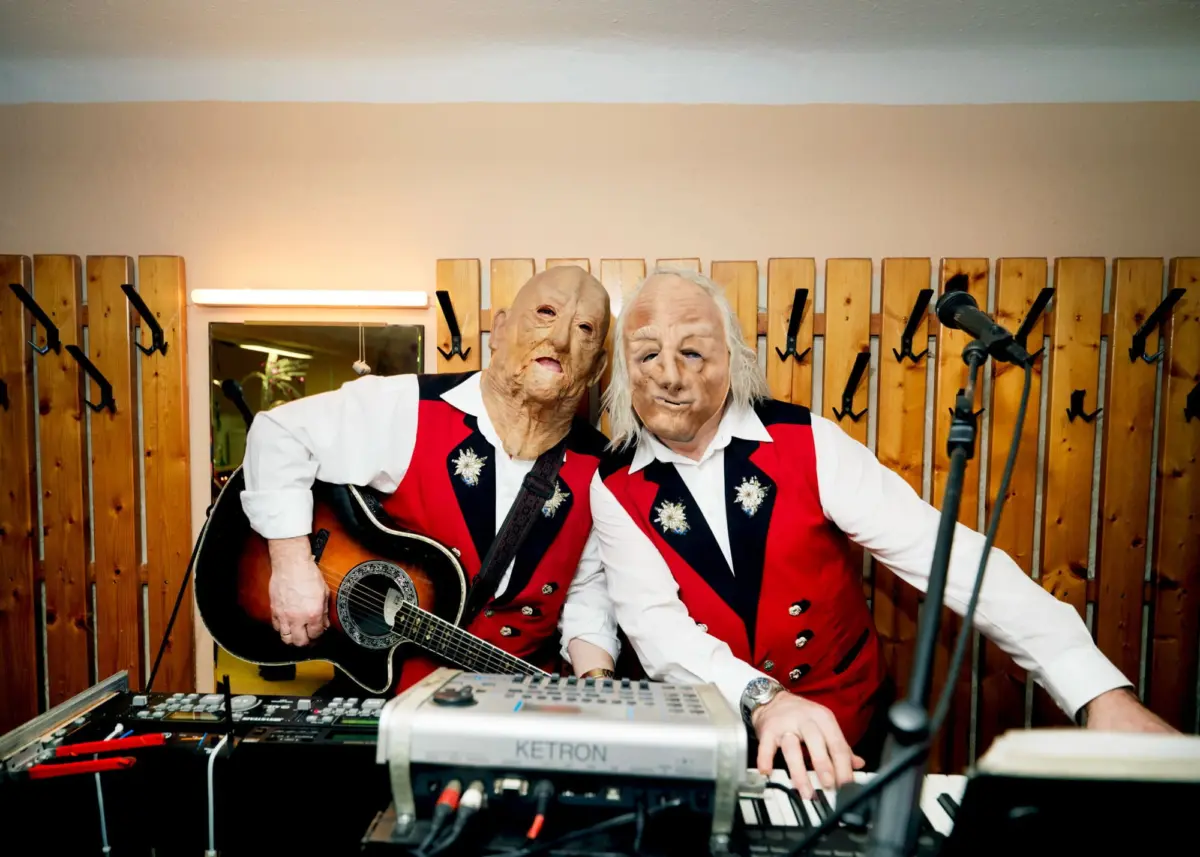Two masked musicians wearing red vests perform with guitar and keyboard in a studio with wooden screen panels and a microphone.