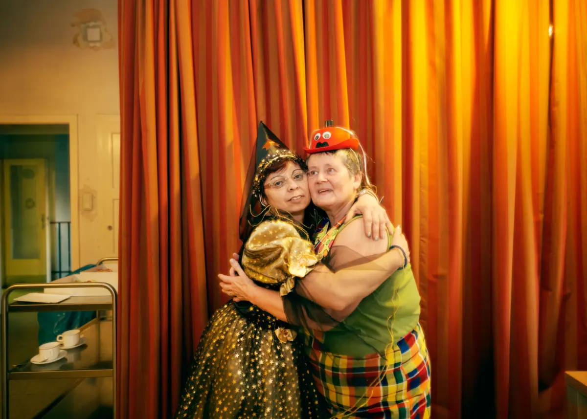 Two people in festive costumes hug in front of orange vertical curtains.