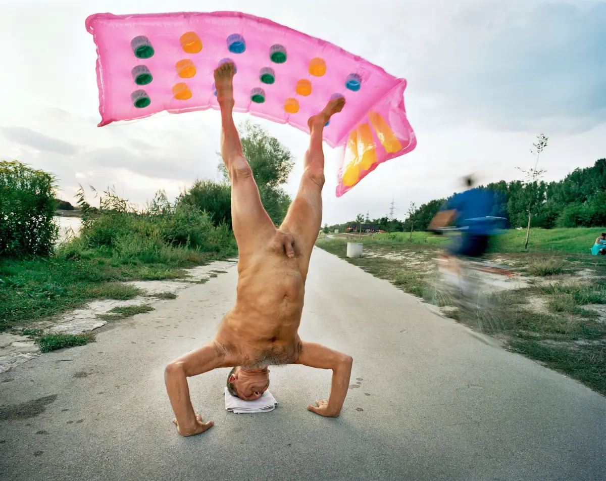 Person performing a handstand on a paved path, holding a pink inflatable pool raft overhead.