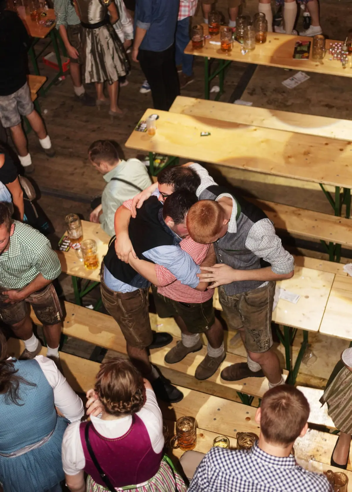 Group of men in lederhosen hugging on wooden benches at a beer hall festival, with beer mugs on tables.