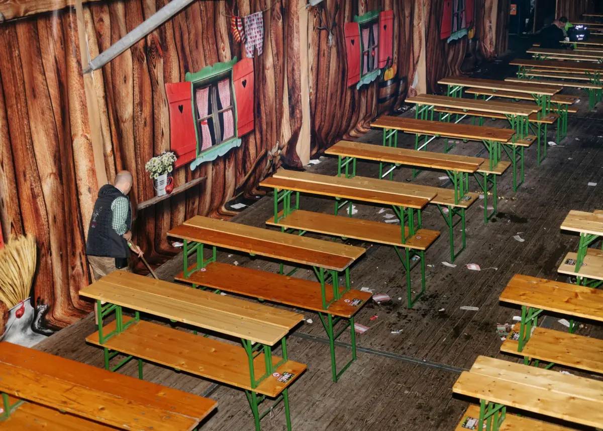 Janitor sweeping wooden floor beside rows of picnic benches in a festival tent with log-wall mural.