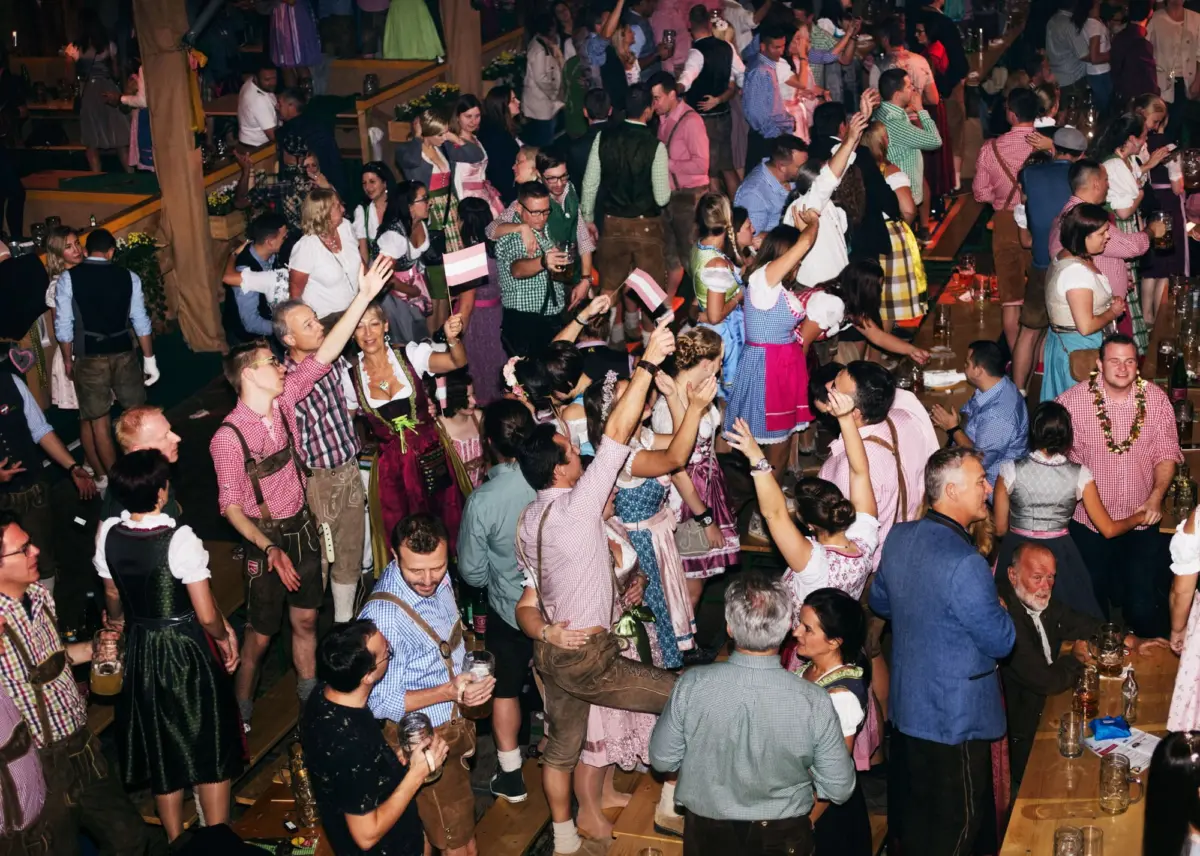 Crowded beer hall with people in dirndls and lederhosen dancing and toasting around long wooden tables.